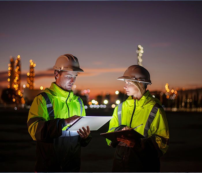 A male and female in a reflective jacket and white hard hats working at an industrial site at night.